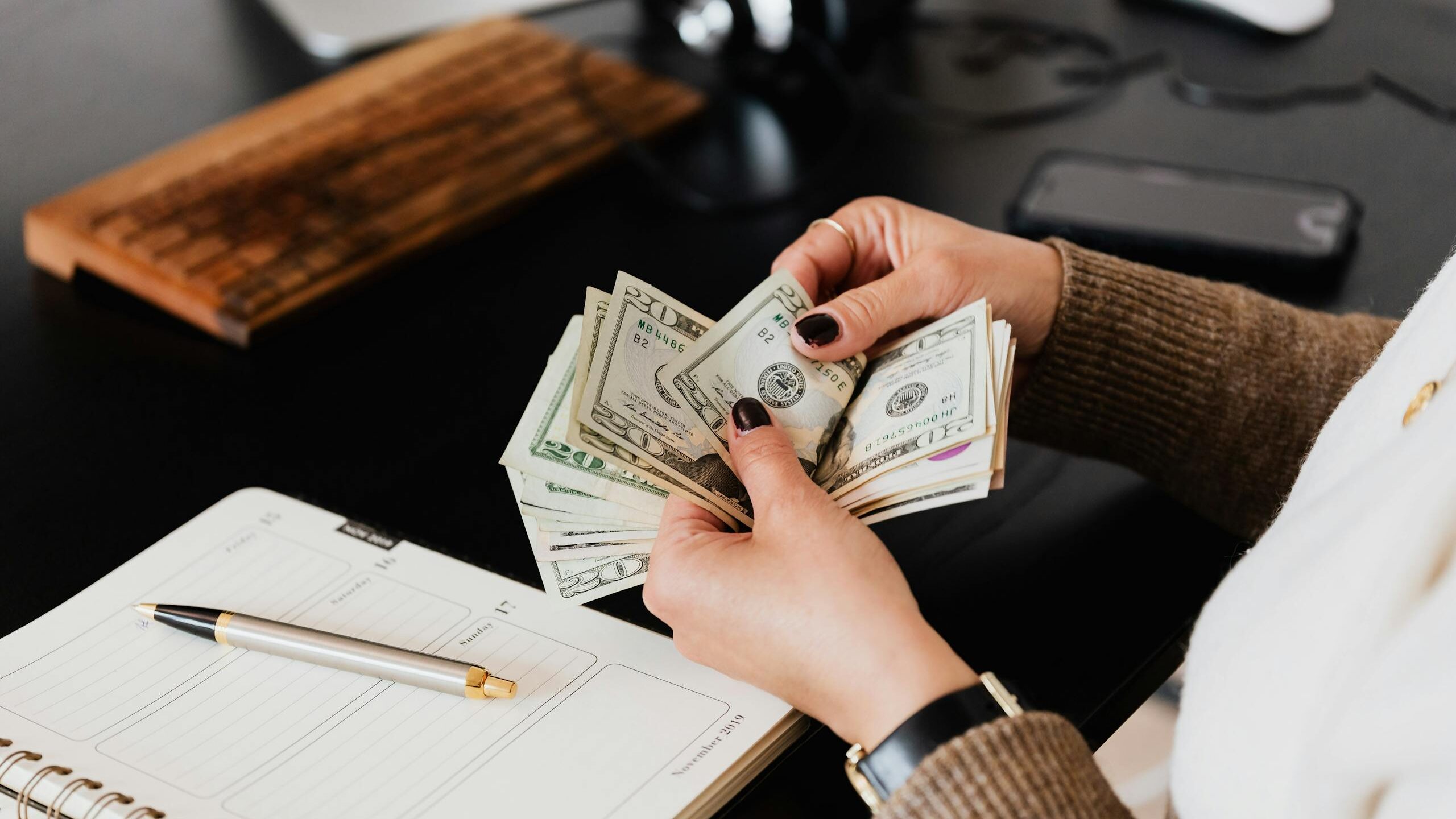 Unrecognizable elegant female in sweater counting dollar bills while sitting at wooden table with planner and pen